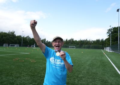 A man in a blue Carr Gomm shirt is showing off his medal with a fist in the air.