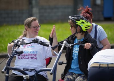 Two one, one in a white Carr Gomm shirt and one in a blue Carr Gomm shirt, are on a joint bicycle. They are high-fiving.