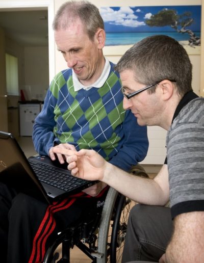 Two individuals, one using a wheelchair, are looking at a laptop together. The setting appears to be a home or office, with a landscape painting in the background.