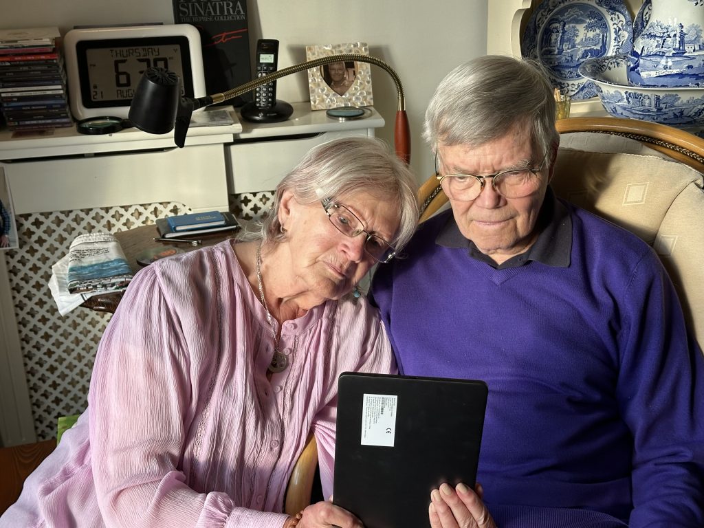 Digital Inclusion Jeff & Marj Two elderly individuals sitting closely on a sofa, looking at a tablet together. The room is decorated with books and traditional ornaments, including a display of blue and white ceramic plates.
