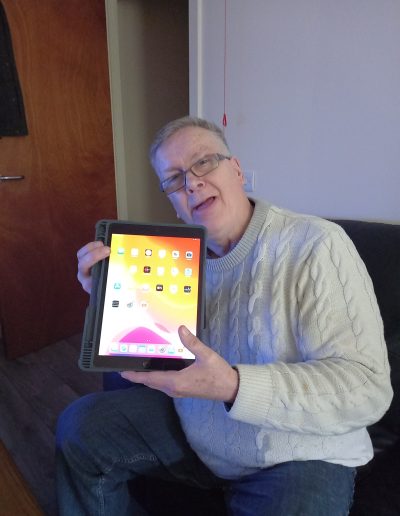 two individuals, one using a wheelchair, are looking at a laptop together. The setting appears to be a home or office, with a landscape painting in the background.