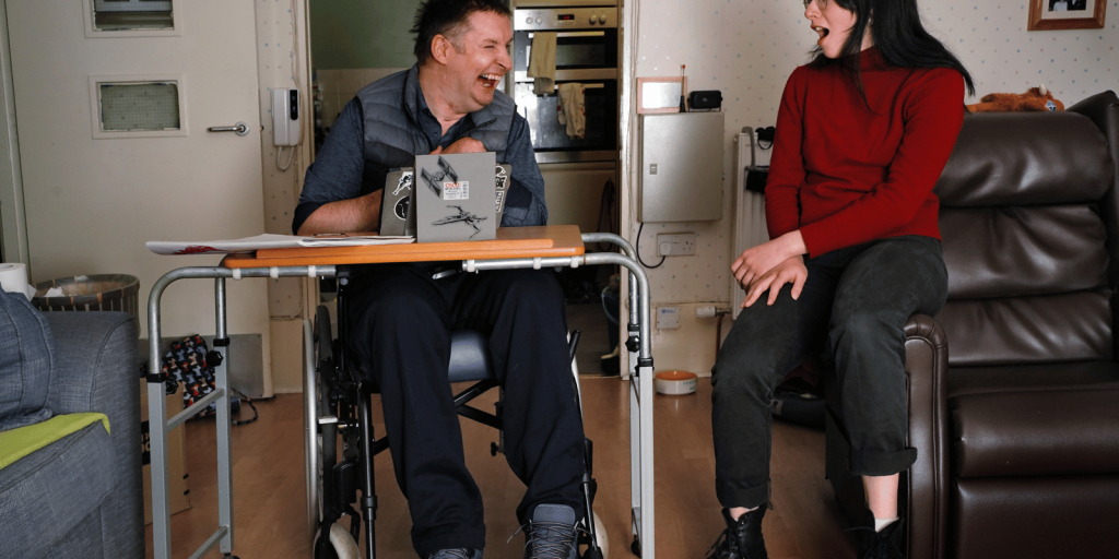 Two people happily conversing in a living room, one seated in a wheelchair and the other on a couch, with a small table between them.