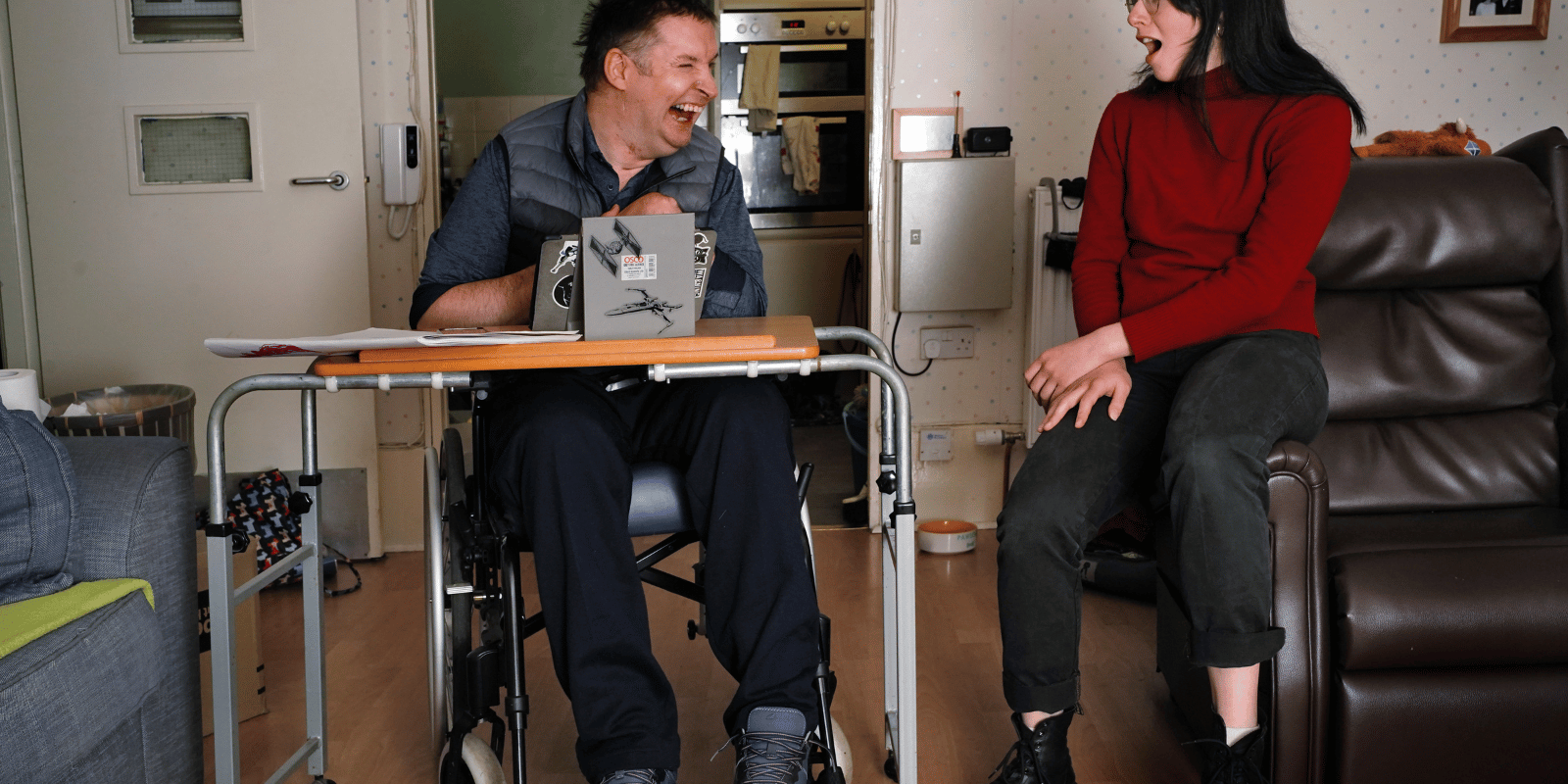 A man in a wheelchair with an ipad on a table smiling and laughing with a young woman sat on couch next to him.