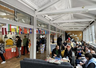 Carr Gomm Glasgow Forum - Inside a community hall with wooden beams on the ceiling and a row of flag bunting hanging in the middle. People are sitting on chairs, chatting. On the left, a table with food can be seen.