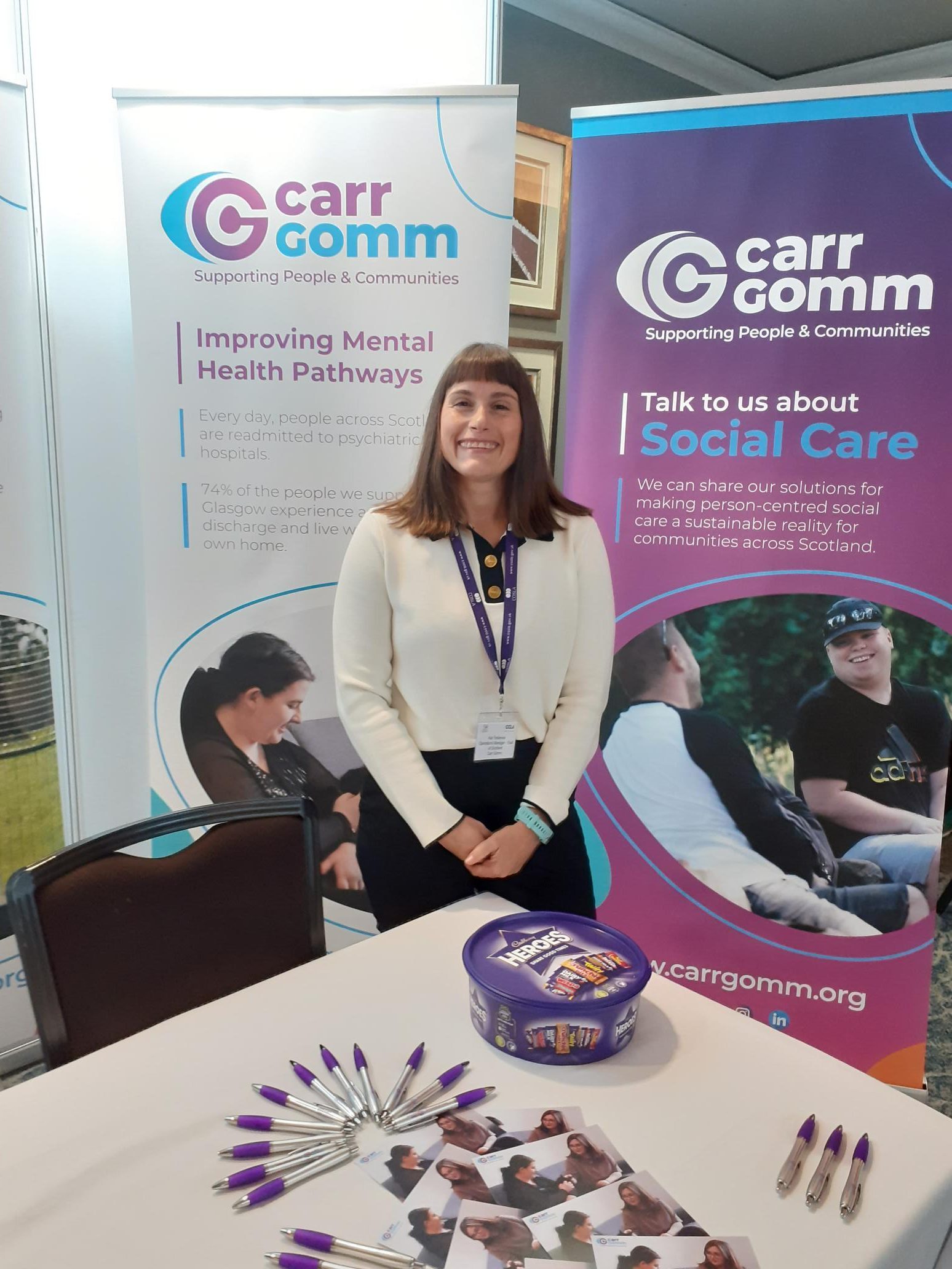 A woman is smiling at the camera. She is surrounded by Carr Gomm pop-up banners and there is a table with sweets and Carr Gomm branded items in front of her at the COSLA conference.