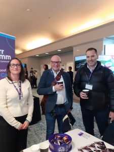 Two men and one women are standing smiling at the camera. They are in a conference room with various stalls around them.