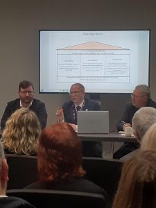 Three men sit at a desk presenting to a group of people.