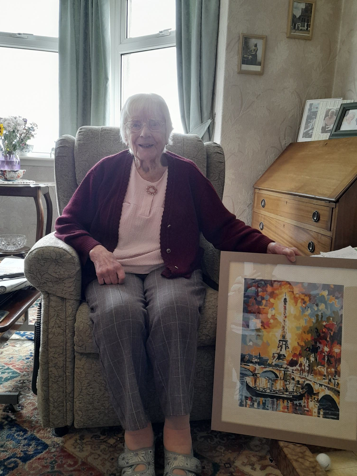 An older woman sitting on an armchair in a living room environment. She has a framed painting by her feet.