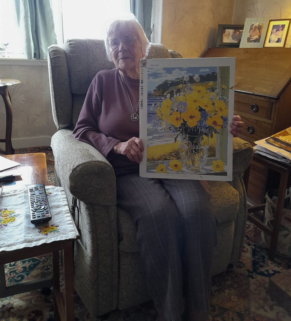 A woman (Katherine) sitting in an armchair holding a large print of Van Gogh's "Sunflowers" painting. The room is decorated with floral patterns and traditional furniture.