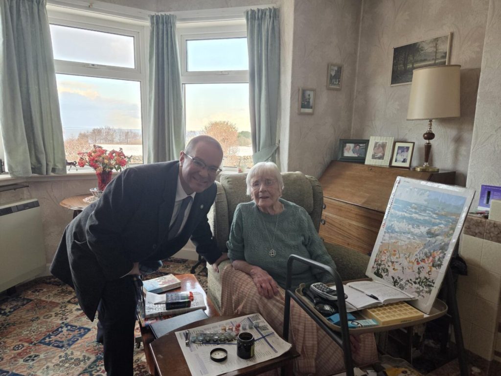 An indoor scene featuring a person in a suit bending slightly with a smile alongside an elderly individual seated in a chair surrounded by paintings, in a cozy room with vintage decor.