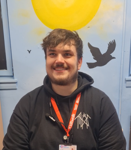 A man in a black hoodie and 'Kiltwalk' lanyard sits in front of a blue wall with a sun and bird painted on it. He is smiling at the camera.