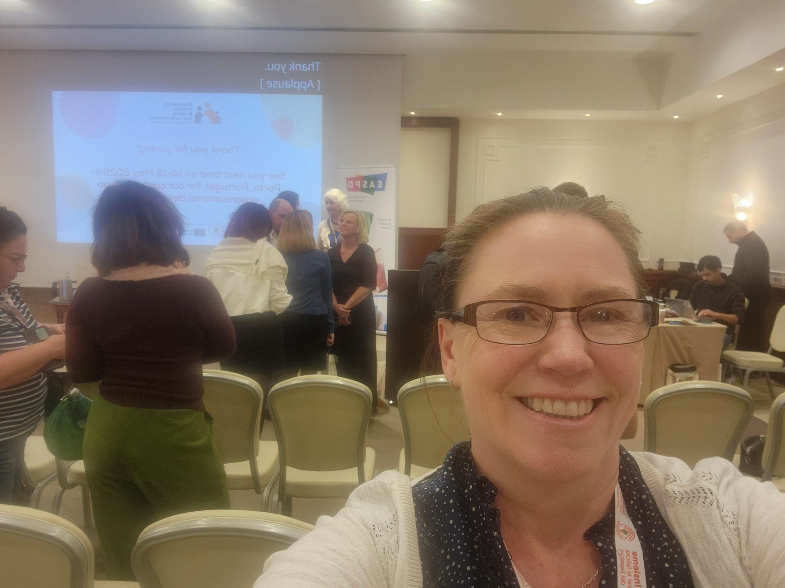 A woman is taking a selfie, she is smiling at the camera. In the background is a conference setting with chairs and a projector. Taken at the EASPD Conference