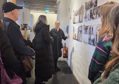 A group of people are listening to a women talk about art that is on the wall in front of them.