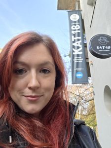 A woman smiles at the camera. A big sign that reads 'Kat 18' can be seen in the background.