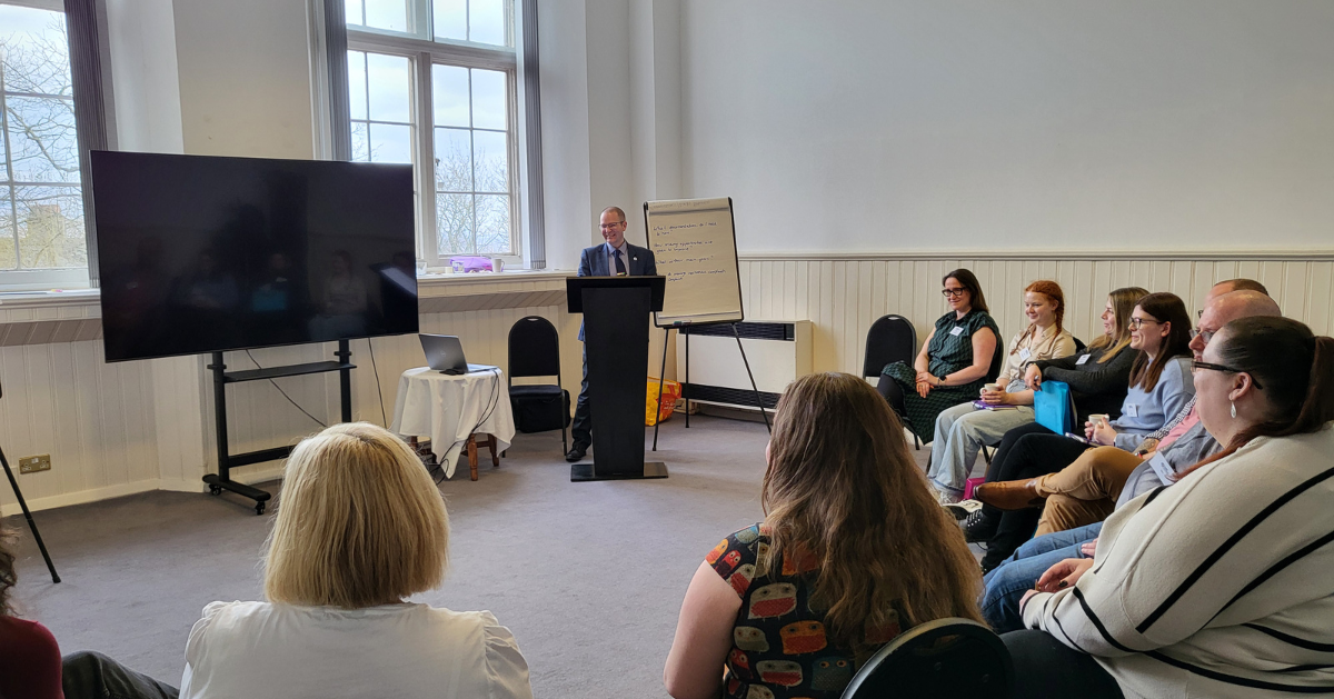 A group of students sit in a semi circle as a man presents from a podium.