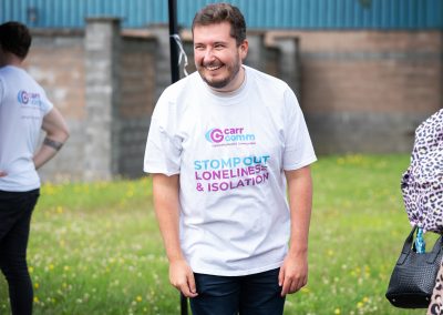 A man (Sandy Hunter, Operations Manager ) is wearing a Carr Gomm t-shirt and smiling at an outdoor event.