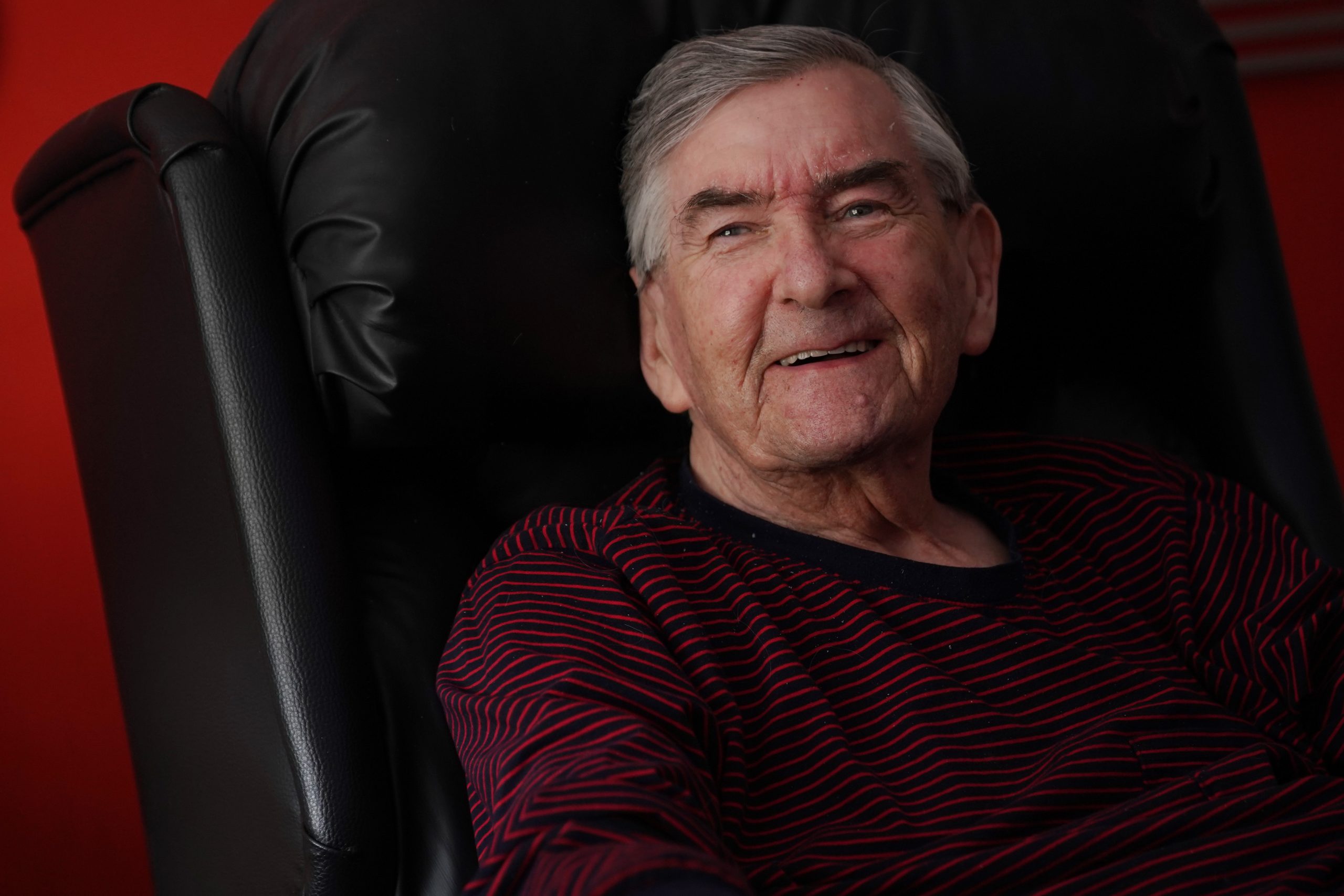 An older man sits in an armchair smiling