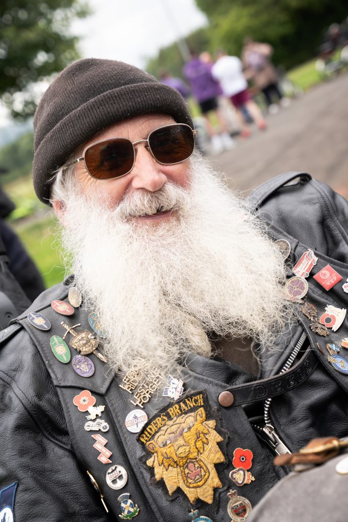 A man (John) wearing sunglasses and a beret, smiling and dressed in a leather jacket adorned with various pins and patches, including a prominent 'Hog's Ranch' patch. | Carr Gomm Physical Disabilities