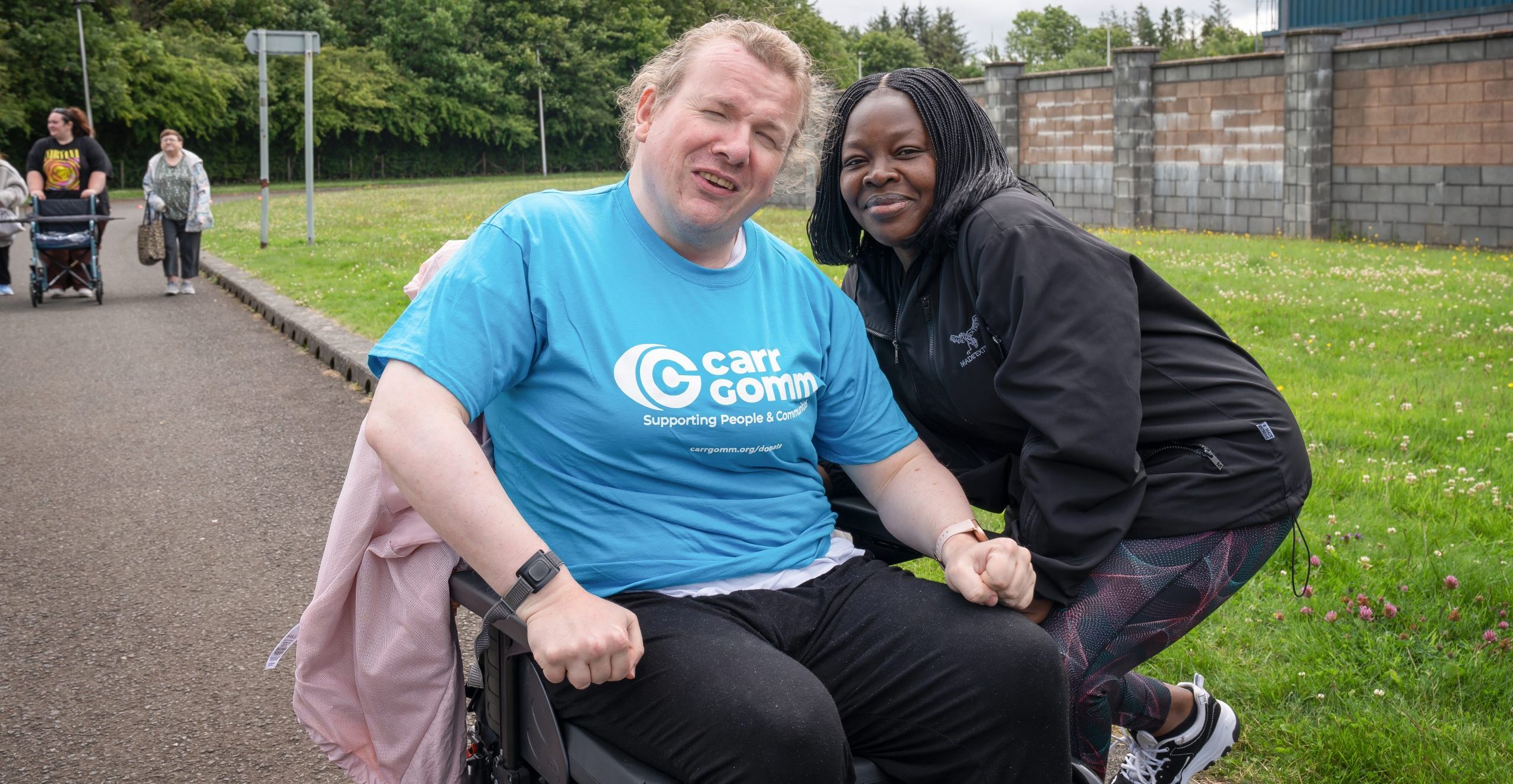 Two women, one in a wheelchair wearing a Carr Gomm t-shirt and the other standing beside them in a black jacket, smiling at the camera outdoors. | Carr Gomm Physical Disabilties