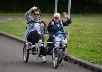 Two women ride on an adaptive bicyle while waving and smiling. One appears to be a Support Practitioner