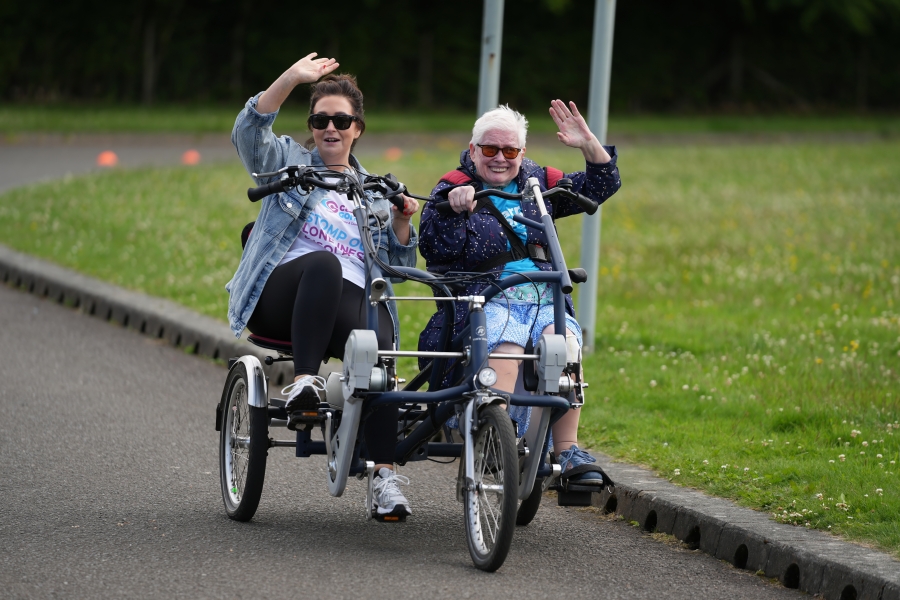 Two women ride on an adaptive bicyle while waving and smiling.