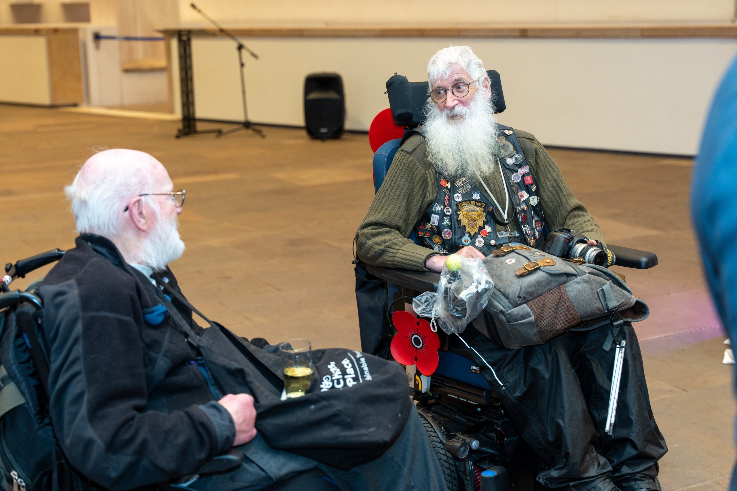 Two individuals in wheelchairs wearing vests decorated with badges and poppies are conversing in an indoor setting.