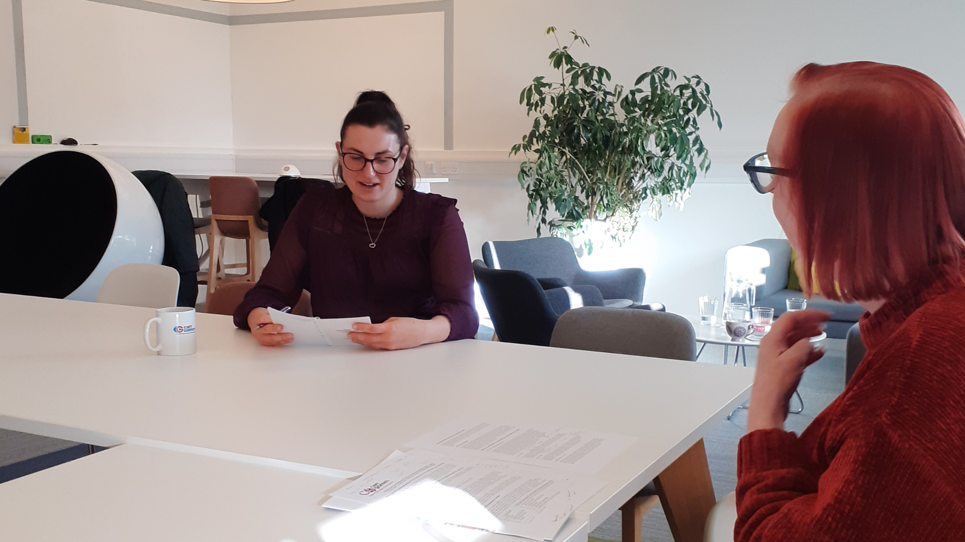 Two woman are conversing. They are sitting at a big white table with papers and a plant in the background.