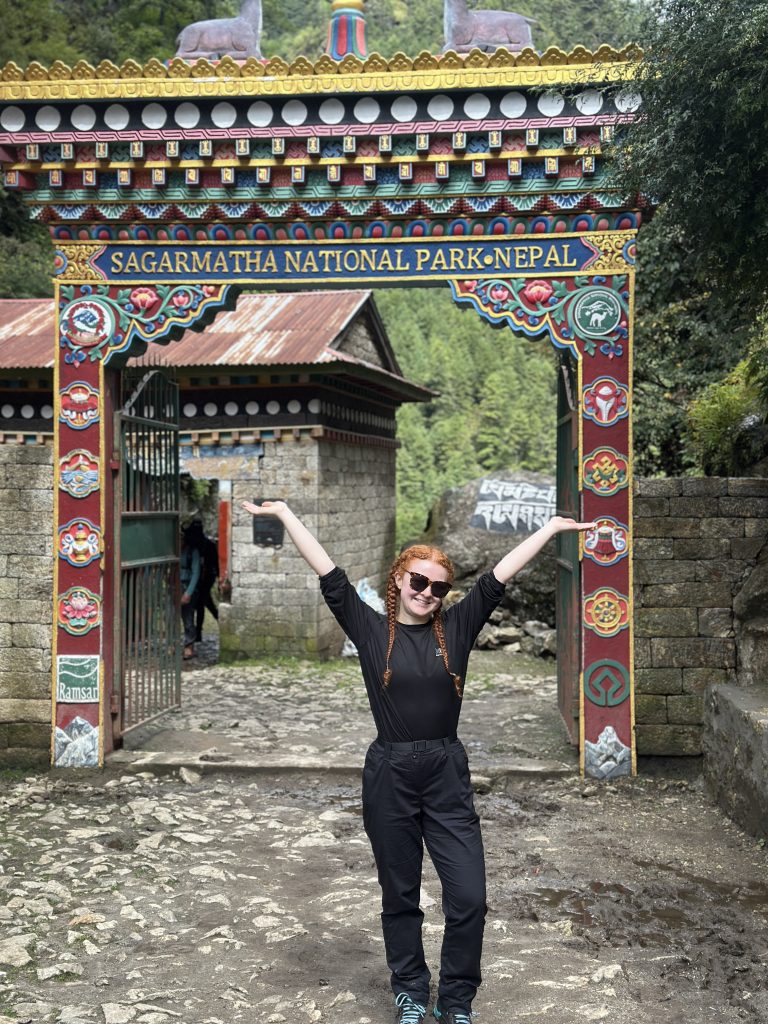 A young woman in hiking clothes poses triumphantly at the entrace to a national park in Tibet.