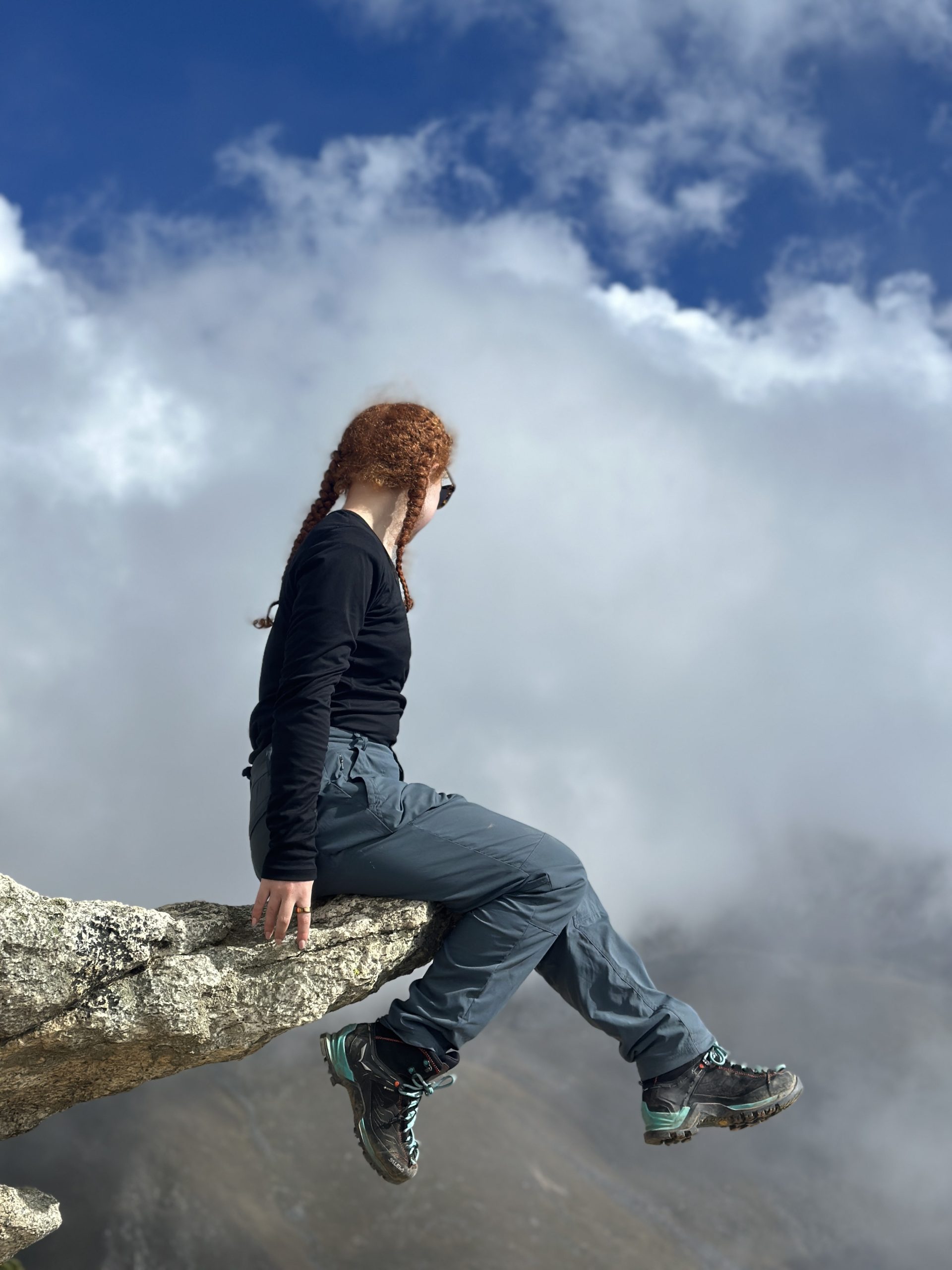 Social Care Careers Daisy A young woman in hiking clothes sits on a cliff edge amongst the clouds.