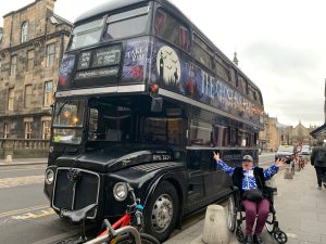 A women in a wheelchair wearing pink leggings has her arms in the air in front of a Ghost tour double decker bus.