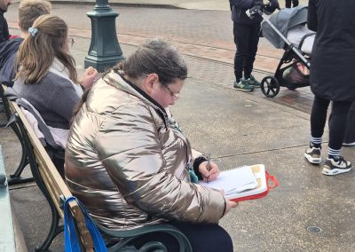 A woman (Edina) sits on a park bench drawing on a sketchbook. She is in a park with lots of people around.
