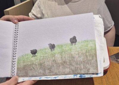 A feminine hand holds up a drawing of black sheep in a field in front of a woman sitting at a cafe booth