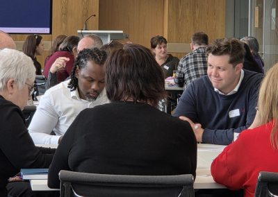 In the foreground, 5 people are sat around a table focusing on writing something. In the background, there are more people sat around a table in a large room.