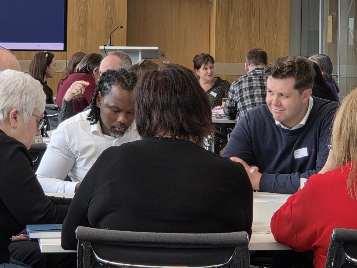 In the foreground, 5 people are sat around a table focusing on writing something. In the background, there are more people sat around a table in a large room.