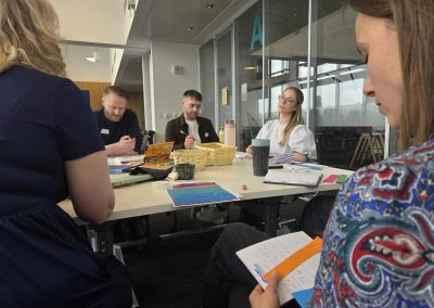 Five people sat around a table, some of them looking at a piece of paper on the table and one looking at another person who is speaking.