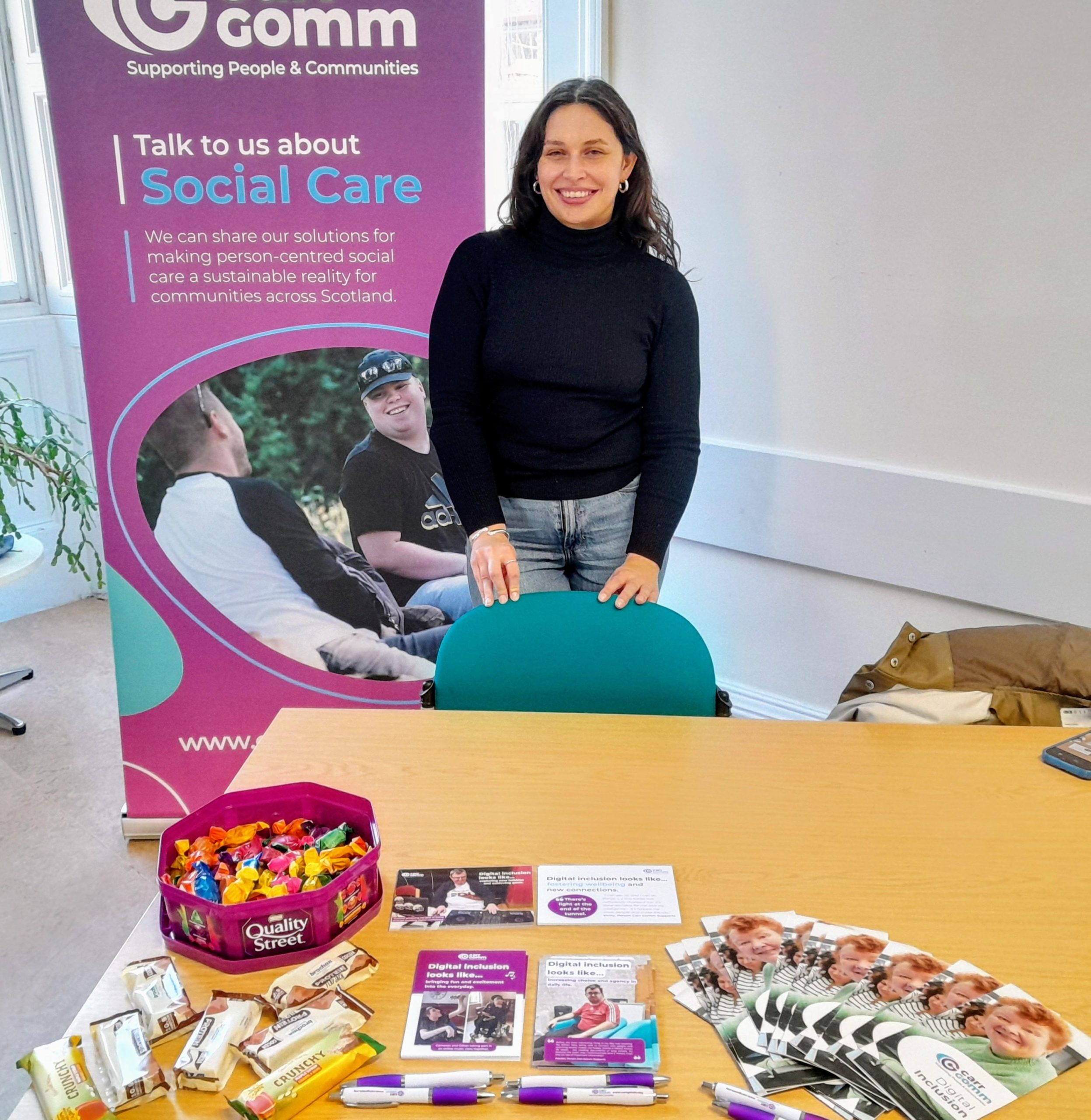 World Social Work Day Woman in dark top and jeans stands in front of Carr Gomm banner and behind table with leaflets and sweeties on it.