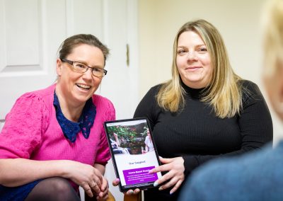Two women (Leanne and Sarah), sit side by side in an office environment holding a tablet.