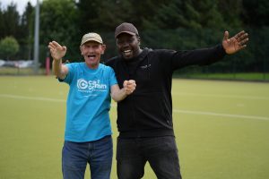 Two men on a sports pitch stood next to each other with arms outstretched and smiling