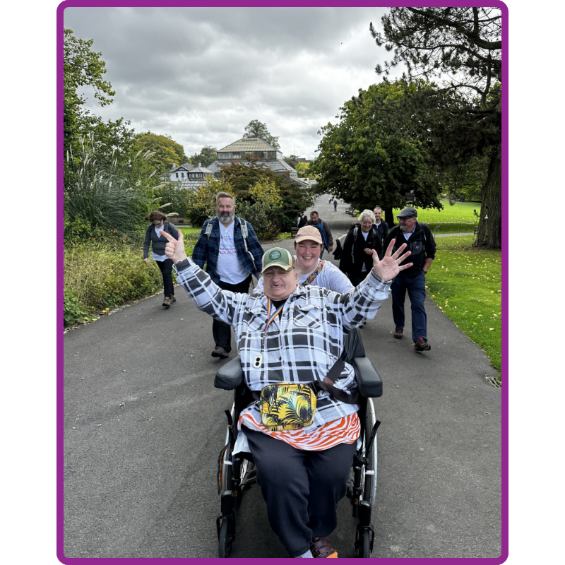 A woman is being pushed in a wheelchair. Shes got her hands up in the air.<br />
They are outside on a path with grass either side and grey clouds in the sky. There are other people walking in the background