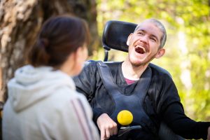 A man (Darryl) who is a wheelchair sits in a wooded area with a wide smile facing a woman wearing a hoodie.