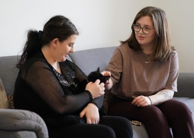 Two young women sit on a sofa in a living room environment. They are stroking a black cat.