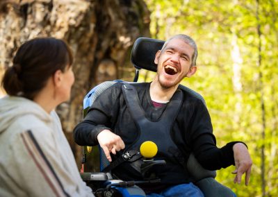A young man (Darryl) smiles widely while looking at a woman who faces away from the camera. Darryl is sitting in a power wheelchair in a sunny woodland.