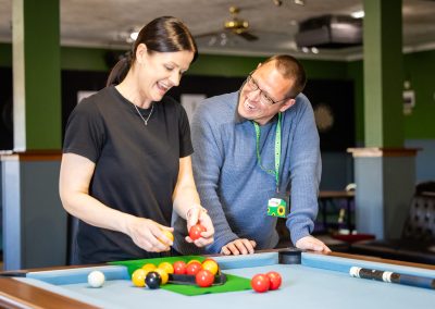 A young woman and a young man are setting up a game of pool in a pool hall. The man wears a sunflower invisible disability lanyard.
