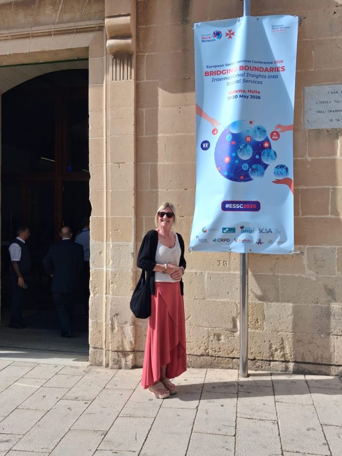 Fiona at the Bridging Boundaries Conference Woman in long skirt and cardigan stood outside in front of banner on a building entitled 'Bridging Boundaries Conference'.