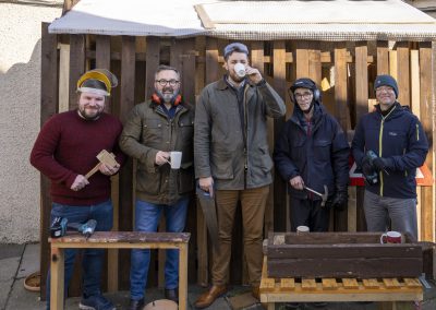 A group of men standing outside a shed holding tools and mugs.