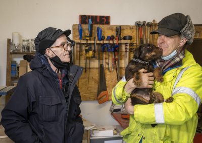 Two men stand an chat in a toolshed. One is wearing a high-vis jacket and holding a small dog.