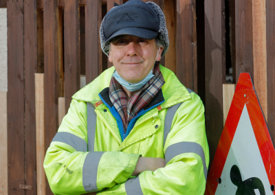 A man (Mark) syands with his arms crossed and smiling. He is wearing a high-vis jacket and wearing a peaked cap.