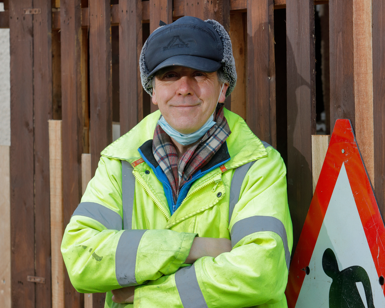 Mark – Mens Sheds blog A man (Mark) syands with his arms crossed and smiling. He is wearing a high-vis jacket and wearing a peaked cap.