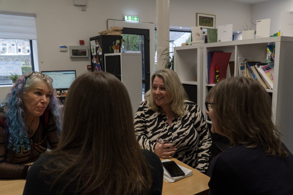 Four women sitting at a desk in an office environment. There is shelving in the background