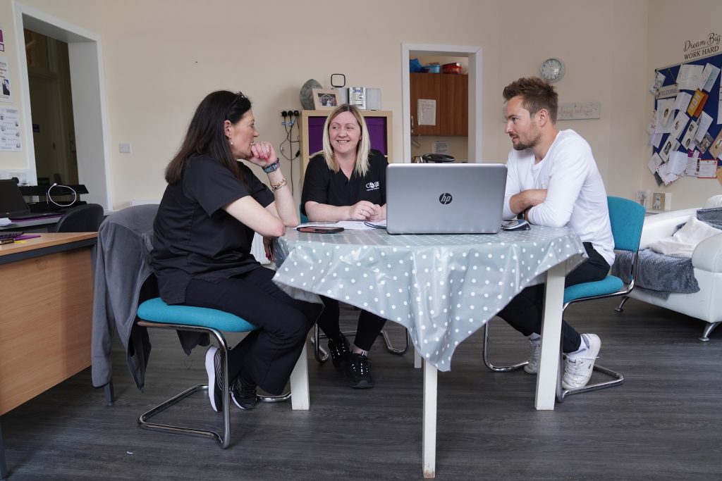 Two women and a man sit around a table having a discussion. On the table there is a light blue polka-dot tablecloth and a laptop. They are in an office environment